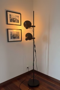 Matte black metal floor lamp with two adjustable spotlight heads, styled in a reading corner next to a vintage armchair